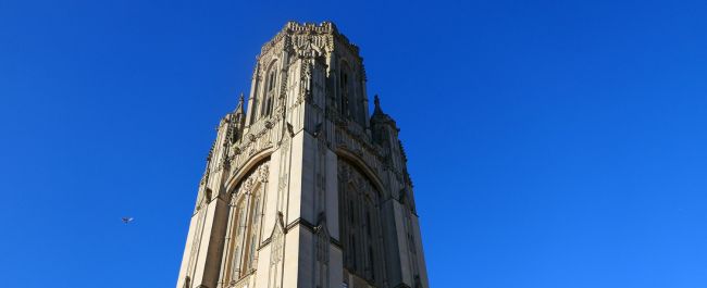 The tower of the Wills Memorial Building against a bright blue sky. A single bird flies past the tower, which is very castle-turret-like and a great feat of gothic architecture. There are gargoyles and other ornamental features that surround beautiful long windows.