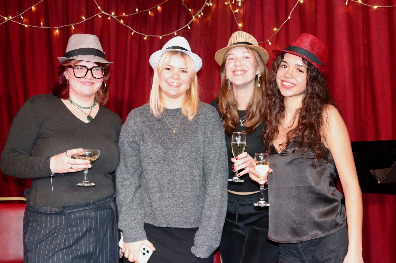 Members of the cast; a group of 4 female students, wearing different coloured hats, on small stage with burgundy velvet curtains in background, festooned with fairy lights.