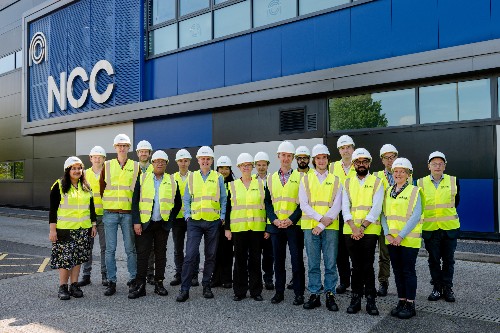 A group of 18 people in hi-viz jackets and white safety helmets standing in front of a building. The letters NCC are on the wall of the building.