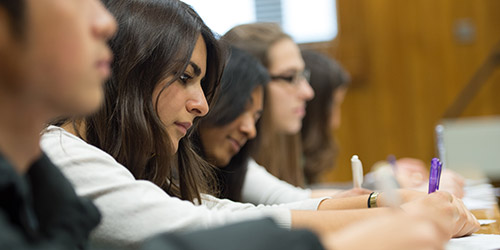 A racially diverse row of students sitting in tiered seating taking notes as they listen attentively to a lecture.