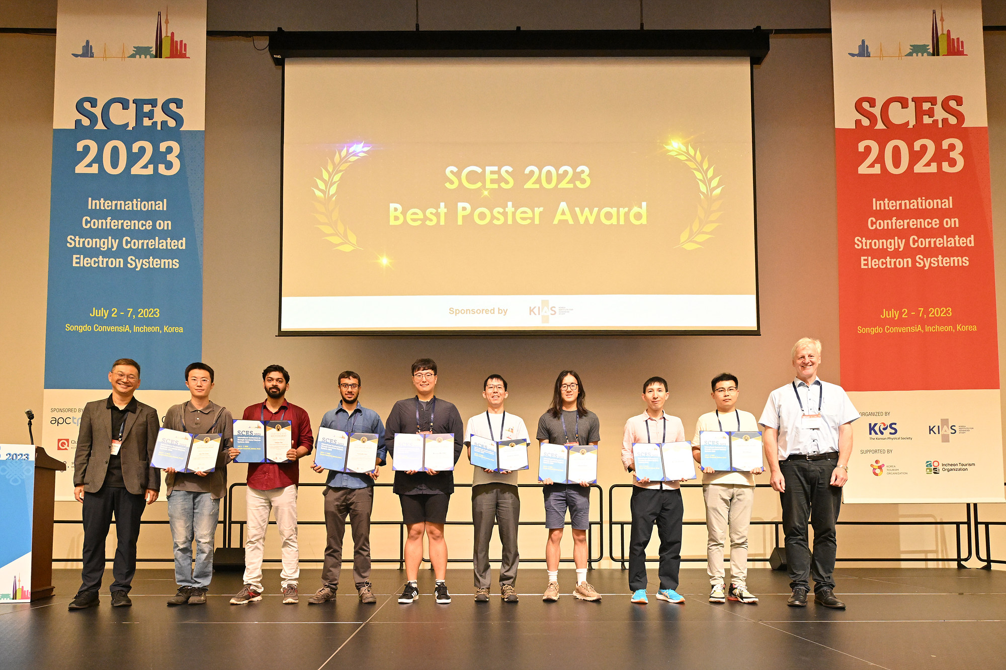 Entrants of the SCES 2023 Poster Prize stand in a row holding certificates. Behind them is a projection which says 'SCES 2023 Best Poster Prize', which is flanked either side by large SCES 2023 banners.