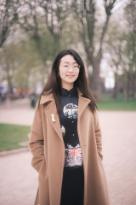 A woman with long dark hair and glasses wearing a brown coat and a black patterned jumper stood in a park