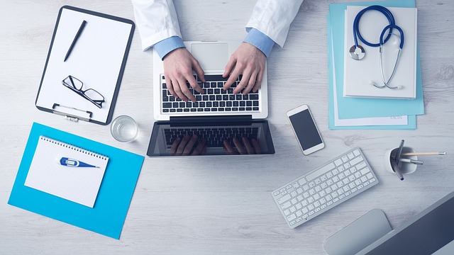 Hands of medic on a laptop with stethoscope, notepad, glasses and mobile phone on table-top