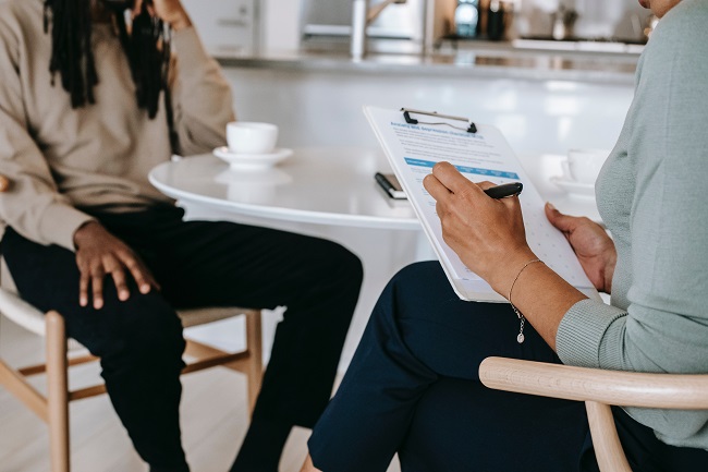 Man seated at table with coffee being interviewed by woman sitting opposite who is completing a questionnaire.