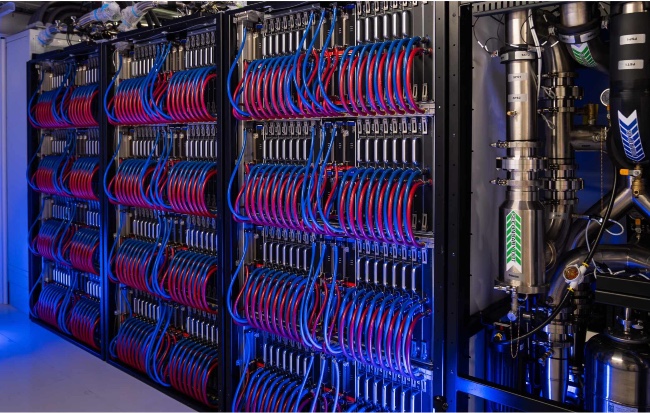An angled photograph of Isamabrd-AI inside the modular data centre. There are blue and red pipes which show the liquid cooling system. There is a blue glow.