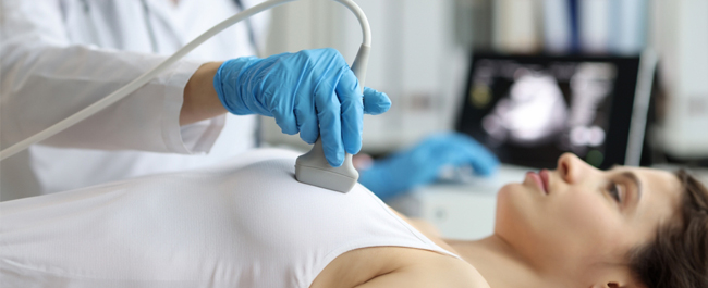 blue latex gloves holding an ultrasound device scan a woman's breast tissue. The woman is wearing a white sleeveless shirt and lying on a bed.
