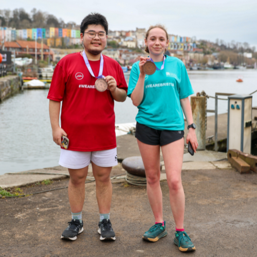 A male and female standing next to each other wearing different coloured #WeAreBristol t-shirts while holding a medal up to the camera