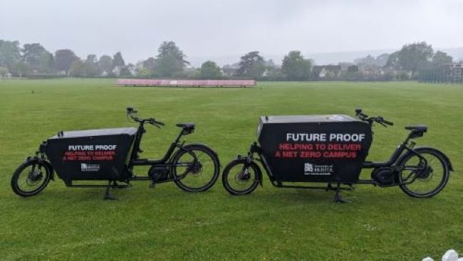 Two University of Bristol Sport Exercise and Health electric black cargo bikes positioned on grass next to cricket pitches. The cargo bikes have a large space at the front for you to place items in for transport and are branded with the text 'Future proof, helping to deliver a net zero campus' on their side.
