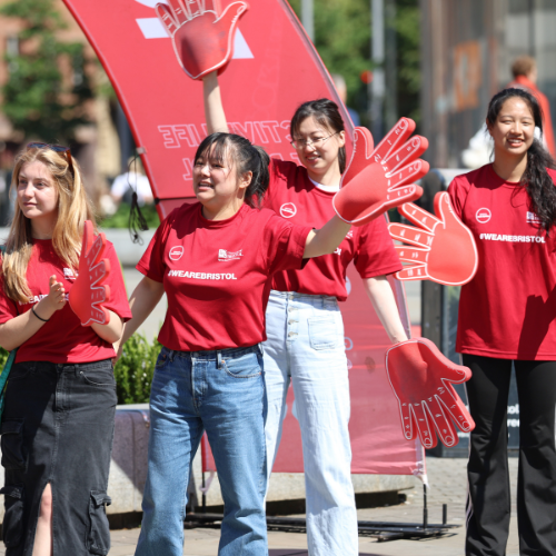 A group of females all wearing red #WeAreBristol t-shirts smiling and waving. Some are wearing red foam hands and waving them around.
