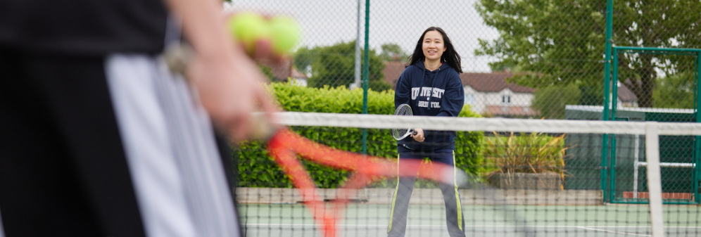 A young adult stands on an outdoor tennis court ready to receive a ball. A hand holds a tennis racket and two tennis balls in the foreground of the photo.