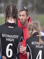In the forefront of the image is the back of 2 school girls. In the background is a man wearing a red jumper coaching these school girls