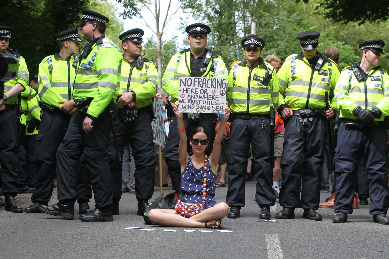Image of a woman sitting crossed legged, holding up a sign against fracking, in front of a line of police