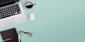 A laptop, notebook, pair of glasses and cup of coffee, all on a desk