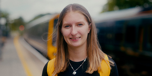 Emmie smiling looking at the camera on a train platform with trains in the background.