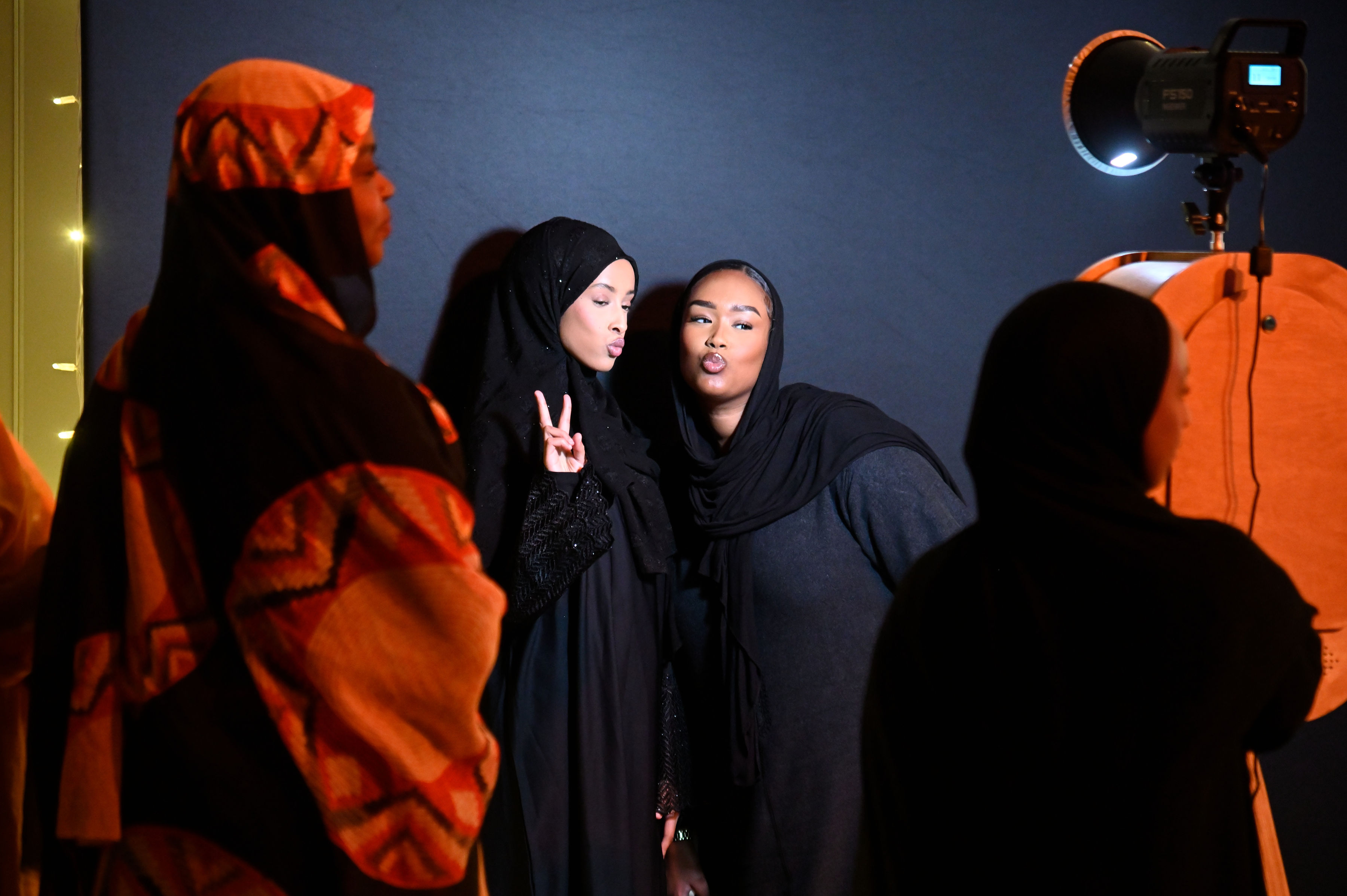 Two women in hijabs pose for a photo against a dark blue backdrop, making peace signs and pouting. A studio light and other guests are visible in the foreground of the photoshoot.