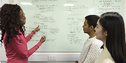 A teacher points at a whiteboard while talking to two students.