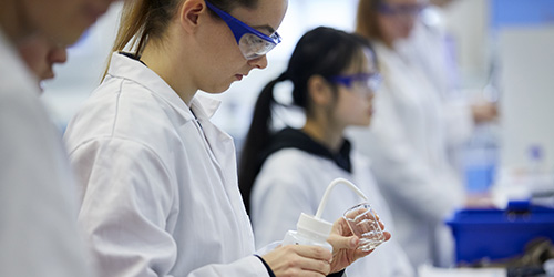 A student dressed in lab coat and goggles focuses on adding liquid to a beaker while other students work around her