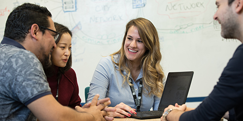 Four people are sat around a laptop having a discussion in a teaching room. 