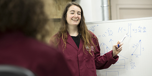 A person wearing a lab coat stands in front of a whiteboard with equations written on.