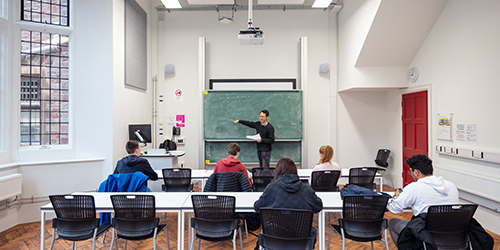 An academic points at an equation on the blackboard at the front of a room with five students looking on