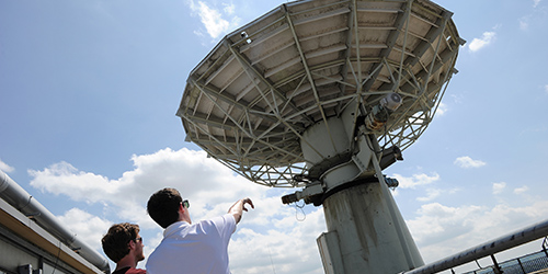 A person points at the a large satellite dish on the roof of a building.