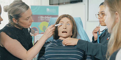 A group of people around a human dummy. One is shining a torch in the eye and one is holding a stethoscope on the chest.