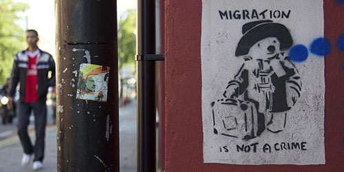A street view with a poster reading 'migration is not a crime' and someone walking past.