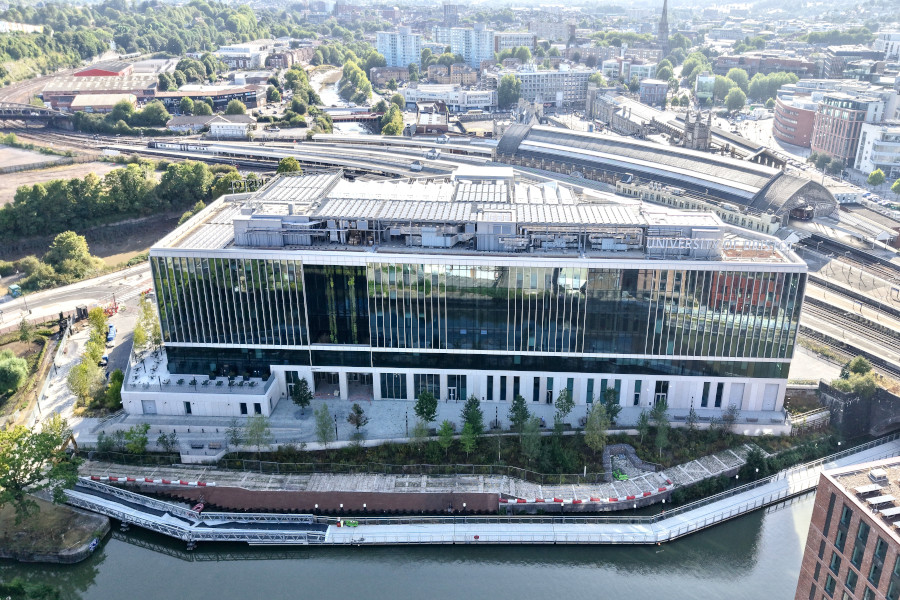 An arial view of a large glass modern building. Infront is a river and behind is a trainline and city scape dispersed with green trees.
