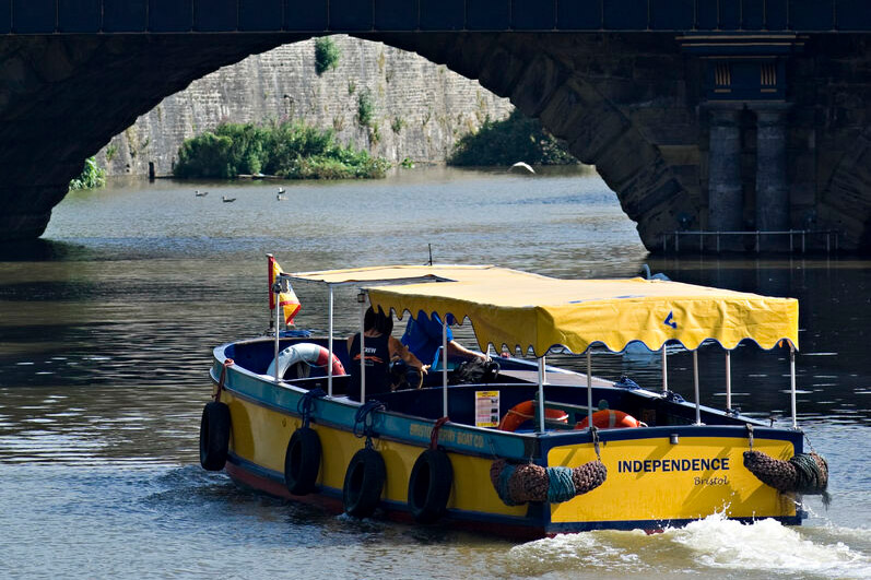 A yellow ferry boat floats along the river Avon heading towards a stone bridge.