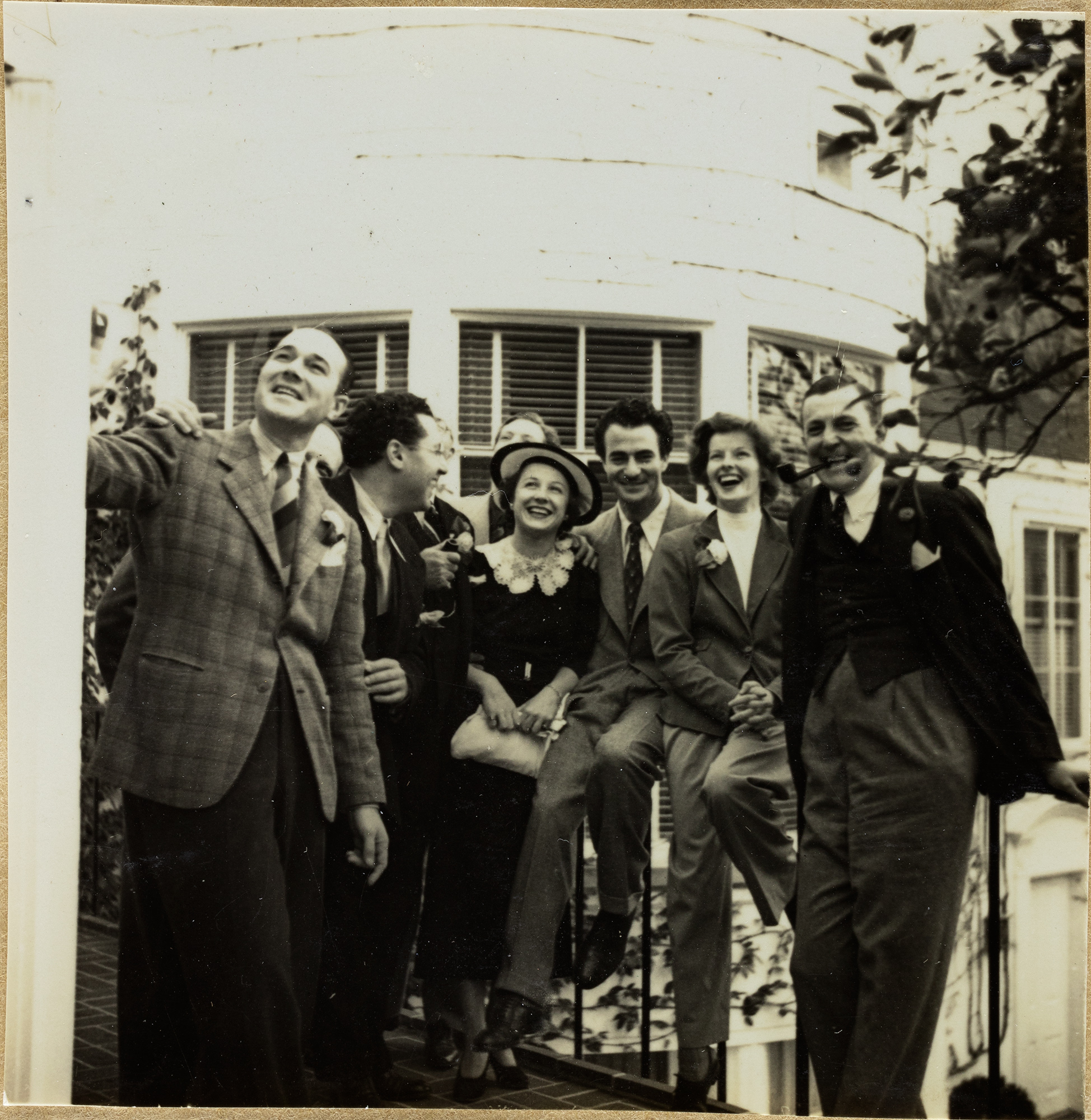 A sepia photograph from the mid-20th century of a group of friends sitting on a rail, the photograph includes Oliver Messel and Katherine H