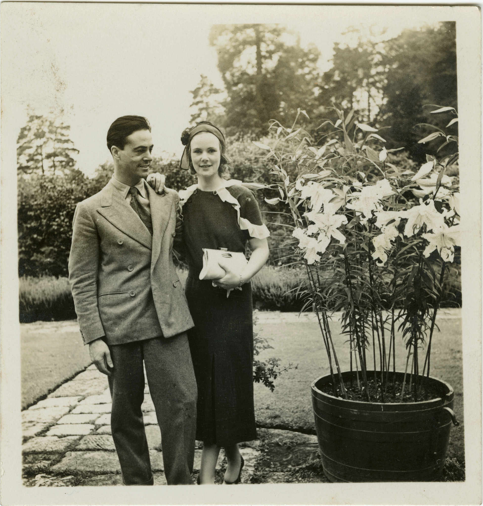 a sepia photograph of oliver messel and a young woman, anne, stood next to a potted plant of flowers