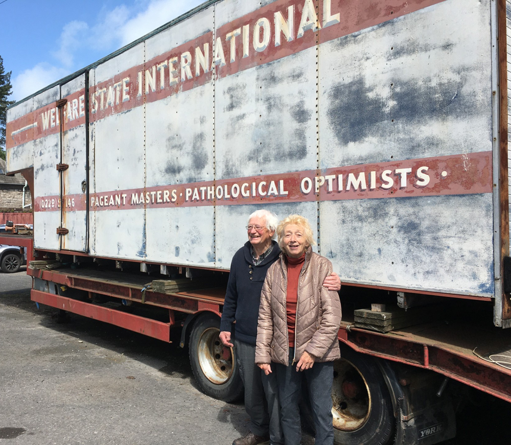 Man and woman standing in front of lorry trailer with the words Pathological Optimists painted on the side