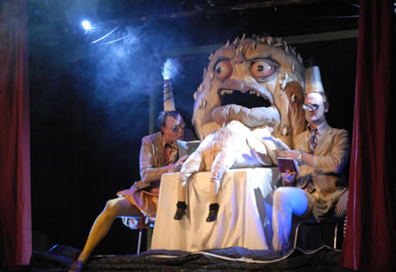 Two men wearing beige suits and sunglasses sit either side of large puppet called Mammon