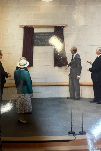 A photograph of five people of different genders stood around a plaque on the wall, with curtains around it. All five people are looking at the plaque and facing away from the camera.