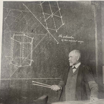 Black and white photograph of a man with short hair and beard, wearing an academic robe and standing in front of a blackboard which has geometric diagrams drawn on it.