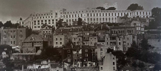 A black and white photograph of several rows of buildings on a hill. A very large multi-storey building stands behind the others, which are made of a darker brick.