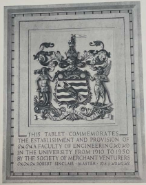 A plaque depicting a coat of arms with a mermaid and a faun stood either side of it, and 'indocilis pauperiam pati' written at the base. Underneath the coat of arms the plaque says 'This tablet commemorates the establishment and provision of a faculty of engineering in the university from 1910 to 1950 by the Society of Merchant Venturers, Robert Sinclair, master, 1953'.