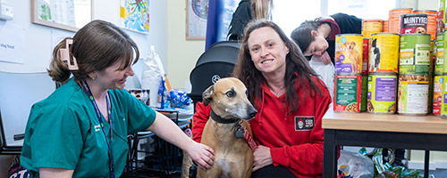 A woman in a red jacket and a woman in scrubs sit with a dog in an indoor setting. Canned food is stacked on a table nearby.