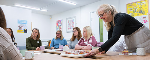 A woman serves a tray of pizza to a group of seated people in a brightly lit room with artwork on the walls.