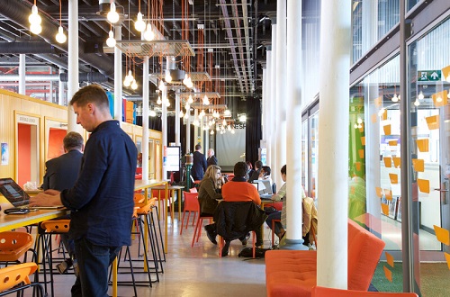 An open industrial space fitted out for comfortable shared working, with pendant lights handing from the high ceiling. There's a person standing typing on a laptop at a standing desk, and a group of people seated on comfortable chairs nearby having a meeting.