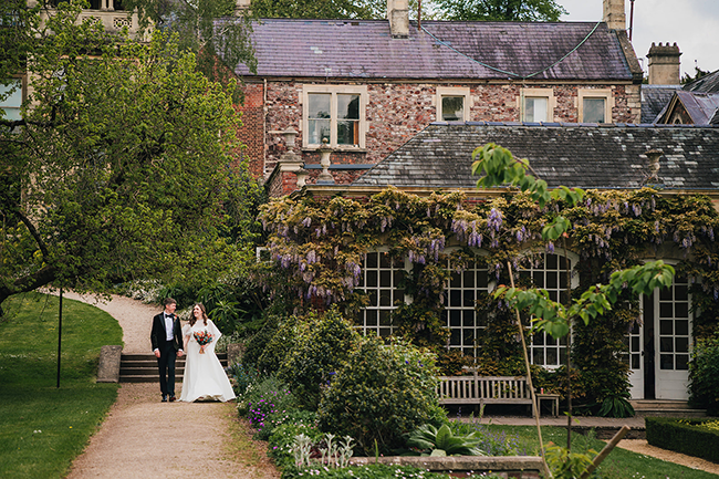 A couple dressed in traditional wedding attire outside an old brick building with flowers on