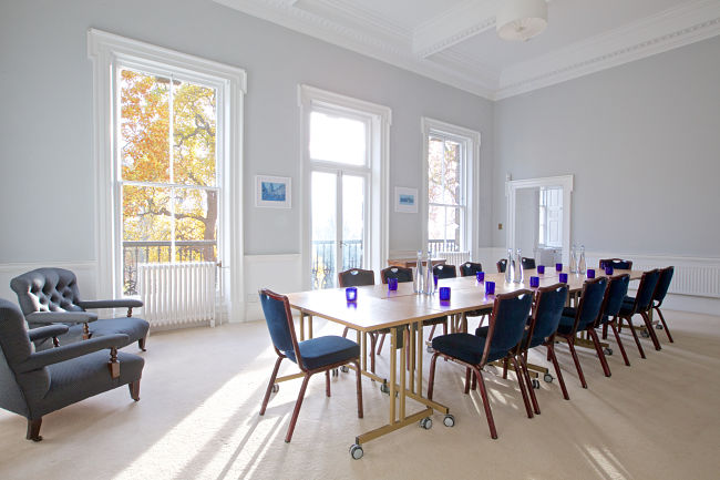 A light spacious room with period features and large windows looking over the gardens. Room setup with boardroom table, glasses and water.