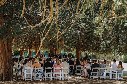 People sat on chairs facing towards a group of people stood up. Behind the people are trees